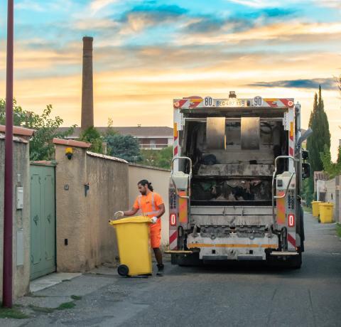 Ceci est une photo où l'on voit des hommes habillés en orange qui ramassent les bacs jaunes pour les mettre dans le camion transporteur. 